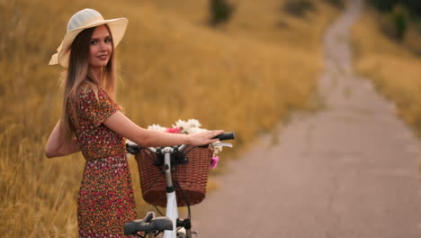 Back-plan-slow-motion:-a-Beautiful-blonde-in-a-dress-with-flowers-in-a-basket-and-a-retro-bike-walks-along-the-road-in-the-summer-field-looking-around-and-smiling-feeling-free.